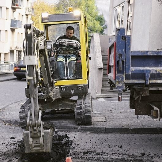 Mini excavator digging a narrow trench beside a road in an urban area.