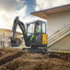Mini excavator digging a trench beside a building under construction.