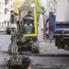 Mini excavator digging a narrow trench beside a road in an urban area.