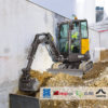 Compact excavator trenching alongside a concrete wall on an industrial construction site.